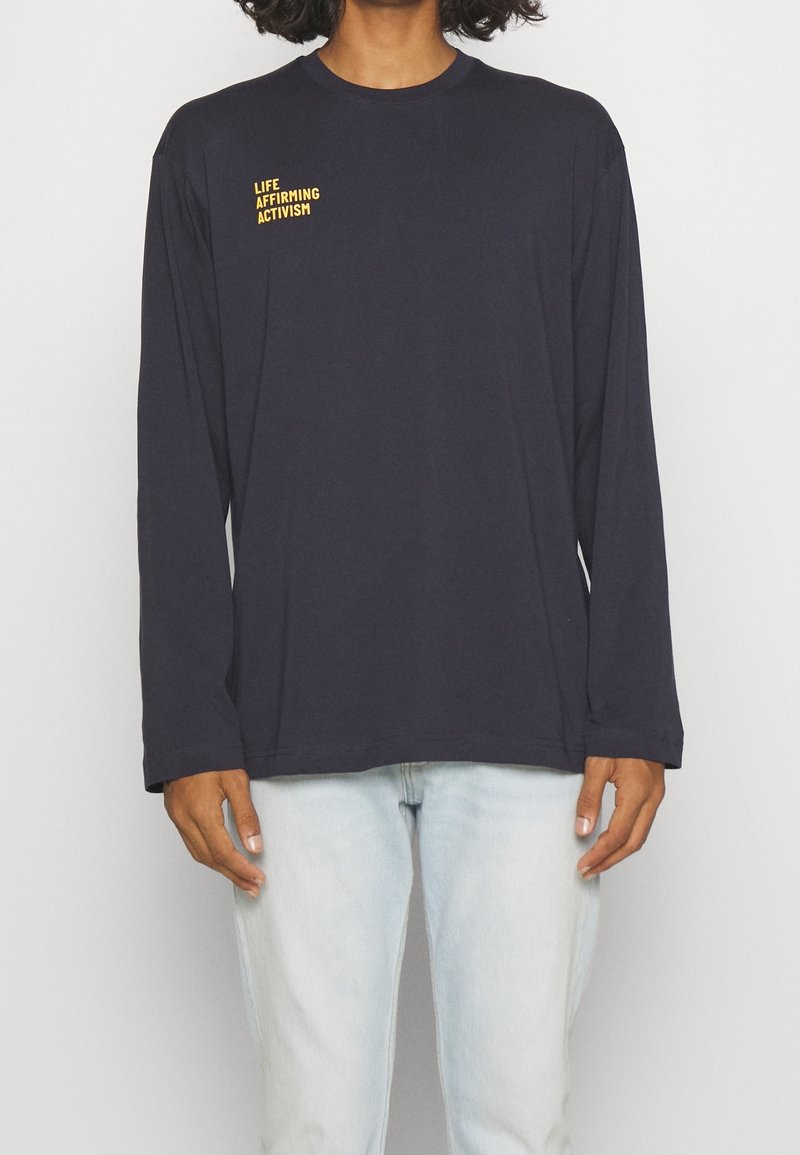 Person som bär en svart långärmad tröja med gul text "LIFE AFFIRMING ACTIVISM" på bröstet, ihop med ljusblå jeans.