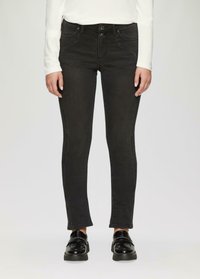 Persoon in zwarte slim-fit jeans, witte lange mouwen top en zwarte glanzende loafers, staande tegen een effen achtergrond.
