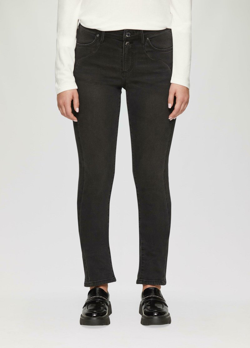 Persoon in zwarte slim-fit jeans, witte lange mouwen top en zwarte glanzende loafers, staande tegen een effen achtergrond.