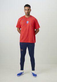 Rdeča kratka majica z Nike logotipom in modrim logotipom PSG, kombinirana z mornarsko modrimi legicami z napisom "PARIS" v zlati barvi. Modri nogometni čevlji dokončajo videz.