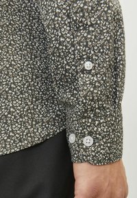 Chemise à manches longues présentant un motif floral sombre en blanc et gris clair, avec des boutons blancs aux poignets et un tissu texturé.