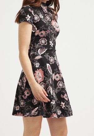 Žena v černých šatech s krátkými rukávy a růžovobílými květinovými vzory stojí před jednobarevným světlým pozadím.