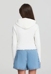 Felpa bianca corta con finitura testurizzata e cappuccio, abbinata a pantaloni corti di jeans blu a vita alta con orlo sfrangiato.