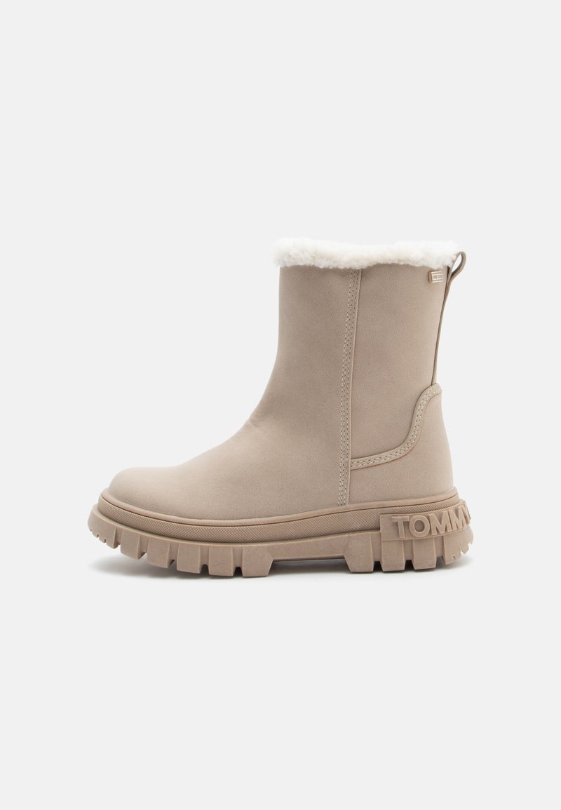 Beige nilkkuri, jossa on pehmeä valkoinen turkisvuori, teksturoitu pohja ja kohokuvioitu logo kannassa. Sillä on sileä ulkopinta ja vetoketju.