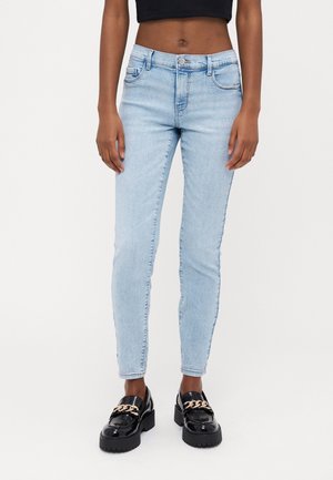 Woman wearing light blue skinny jeans, black cropped top, and black loafers with gold chain detailing, standing on white background.