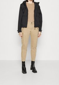 Svart lättviktig jacka med huva, beige fuskskinn cargo-byxor och svarta klackstövlar. Strukturert tyg och funktionella fickor synliga.