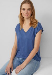 Frau mit hellbraunem Haar, die ein blaues ärmelloses V-Ausschnitt-Top und hellblaue Jeans trägt, sitzt vor einem schlichten grauen Hintergrund.