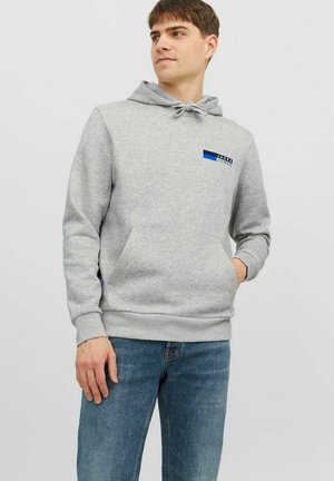 Grauer Kapuzenpullover aus Baumwollmischung mit Kordelzugkapuze, Fronttasche und Logo-Patch in Blau und Schwarz. Modell trägt ihn mit blauen Jeans.