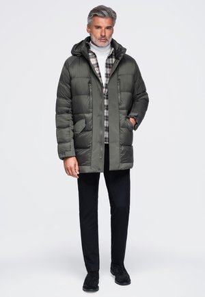Homme portant une doudoune à capuche vert foncé par-dessus une chemise à carreaux et un col roulé blanc, pantalon noir et chaussures noires, debout sur fond blanc.