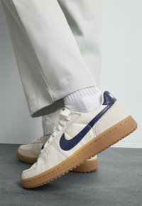 Inimene kannab beežide Nike jalatsitega, millel on tumesinine swoosh, valgeid sokke ja heledat värvi pükse, seistes hallil põrandal heleda tausta ees.