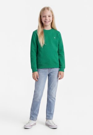 Fată zâmbitoare cu păr lung blond, purtând un hanorac verde, blugi albaștri deschiși și adidași albi, stând pe un fundal alb.