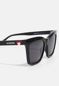 Black oversized sunglasses with gray lenses. Features gold lettering "LOVE MOSCHINO" and a red heart accent on the temple. Smooth plastic frame.