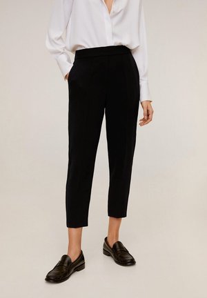 Person wearing black cropped tailored pants, white long-sleeve blouse, and black loafers, standing on light background.