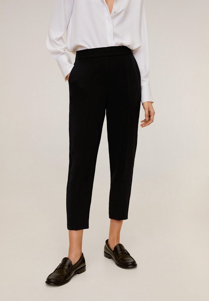 Personne portant un pantalon noir court et ajusté, un chemisier blanc à manches longues, et des mocassins noirs, debout sur un fond clair.
