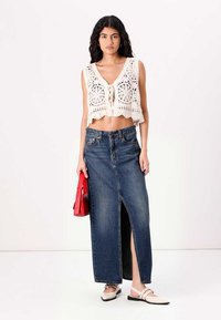 Biele háčkované crop top s viazaním vpredu, skombinovaný s dlhou denimovou sukňou s predným rozparkom. Model drží červenú kabelku a má na sebe ploché topánky.