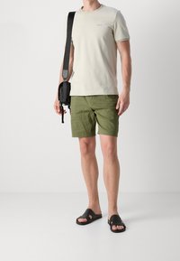 Camicetta grigio chiaro a maniche corte con piccolo logo, pantaloni corti verdi con vestibilità rilassata, ciabatte nere e una borsa a tracolla nera.
