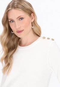 Maglione bianco con scollatura rotonda, impreziosito da bottoni dorati decorativi sulla spalla; il modello ha capelli biondi mossi e indossa orecchini di perle.