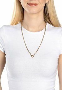 Guld-farve perle halskæde med en cirkulær vedhæng, båret over en tætsiddende hvid t-shirt, der har et tekstureret kædedesign.