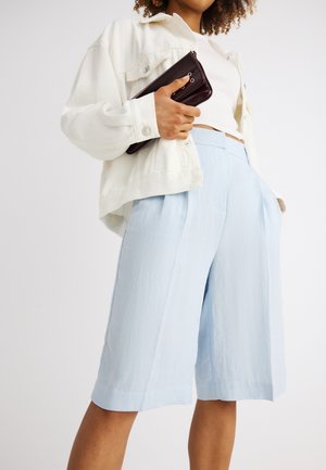 Frau trägt hellblaue, weit geschnittene Shorts bis zum Knie, weißes kurzes Oberteil und weißen, knöpfbaren Blazer, hält eine dunkle Clutch-Tasche.