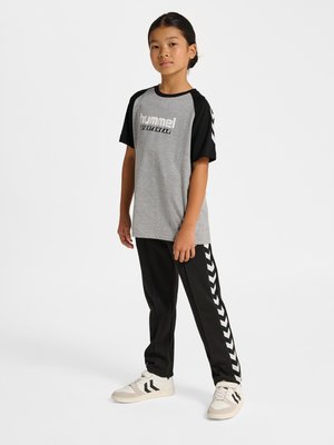 Kind steht und trägt ein graues und schwarzes hummel Sportshirt, schwarze Hose mit weißen Chevron-Streifen und weiße Sneakers.