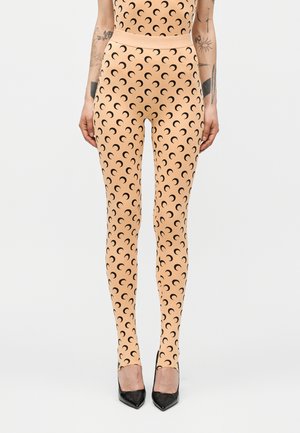 Κολάν σε λεπτή εφαρμογή, χρώματος ροδάκινο, με επαναλαμβανόμενο μαύρο μοτίβο ημισελήνου, φορεμένα με μαύρες μυτερές γόβες, εμφανή τατουάζ στα χέρια.