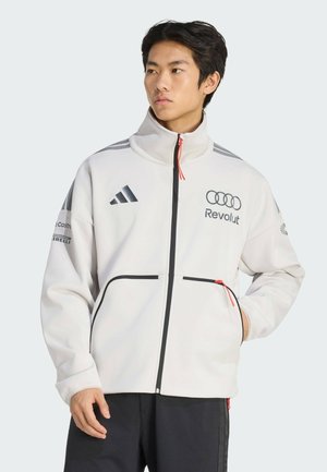 Jeune homme portant une veste blanche zippée avec des détails noirs et rouges et des logos de sponsors, debout avec une main dans la poche, regardant vers la gauche.