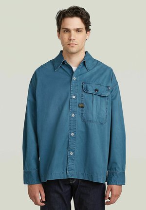 Jonge man draagt een los blauw overhemd met knopen en een grote borstzak en donkere broek, staand tegen een lichtgrijze achtergrond.