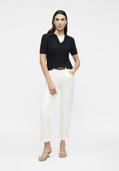 Γυναίκα που στέκεται με το ένα χέρι στην τσέπη, φοράει μαύρο πόλο με κοντά μανίκια, λευκό παντελόνι, καφέ ζώνη και μπεζ σανδάλια με τακούνι.