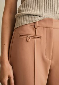 Pantalones beige ajustados con un bolsillo lateral con cierre de botón y una tela suave y texturizada, combinados con una blusa de punto ligera con canalé.