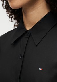 Μαύρο πουκάμισο με μυτερό γιακά, που φέρει μικρό κεντητό λογότυπο σε κόκκινο, λευκό και μπλε στην αριστερή πλευρά. Ομαλή υφή υφάσματος.