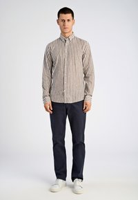 Camisa de manga larga a rayas marrón y blanca con cuello abotonado, combinada con pantalones navy y zapatillas blancas, presentada en un modelo masculino.