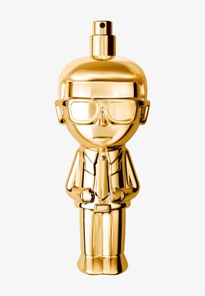 Błyszcząca złota butelka z rozpylaczem w kształcie stylizowanej postaci noszącej okulary, garnitur i krawat, stojącej pionowo na białym tle.