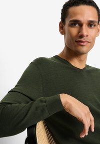 Maglione verde lavorato a maglia con scollo a V, texture liscia e maniche aderenti; modello con la mano appoggiata su una sedia con seduta in paglia intrecciata.