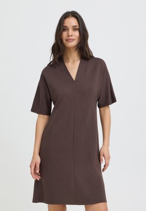 Femme aux longs cheveux foncés portant une robe marron ample à manches courtes et col en V, debout devant un fond clair uni.