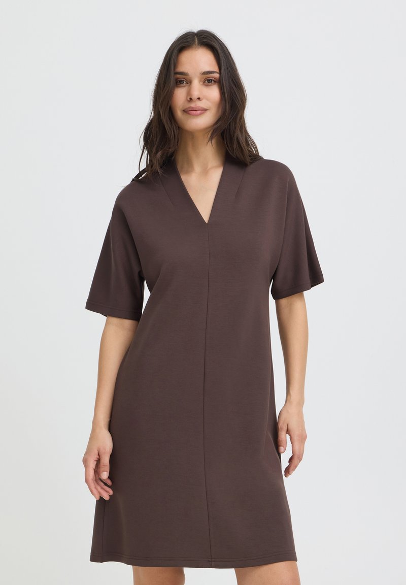 Femme aux longs cheveux foncés portant une robe marron ample à manches courtes et col en V, debout devant un fond clair uni.