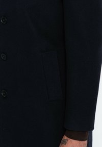 Cappotto di lana blu navy con una texture liscia, dotato di sei bottoni neri, una tasca laterale e una vestibilità sartoriale visibile nei dettagli delle maniche e della tasca.
