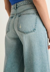 Svetlomodré džínsy s vysokým pásom a rovnými nohavicami, päť vreciek a jemné stehy. Textúrovaná tkanina s drobnými známkami opotrebovania.