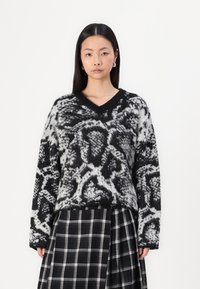 Maglione lavorato a maglia in bianco e nero con motivo, scollo a V, spalle scese e maniche lunghe, abbinato a una gonna a quadri in bianco e nero.