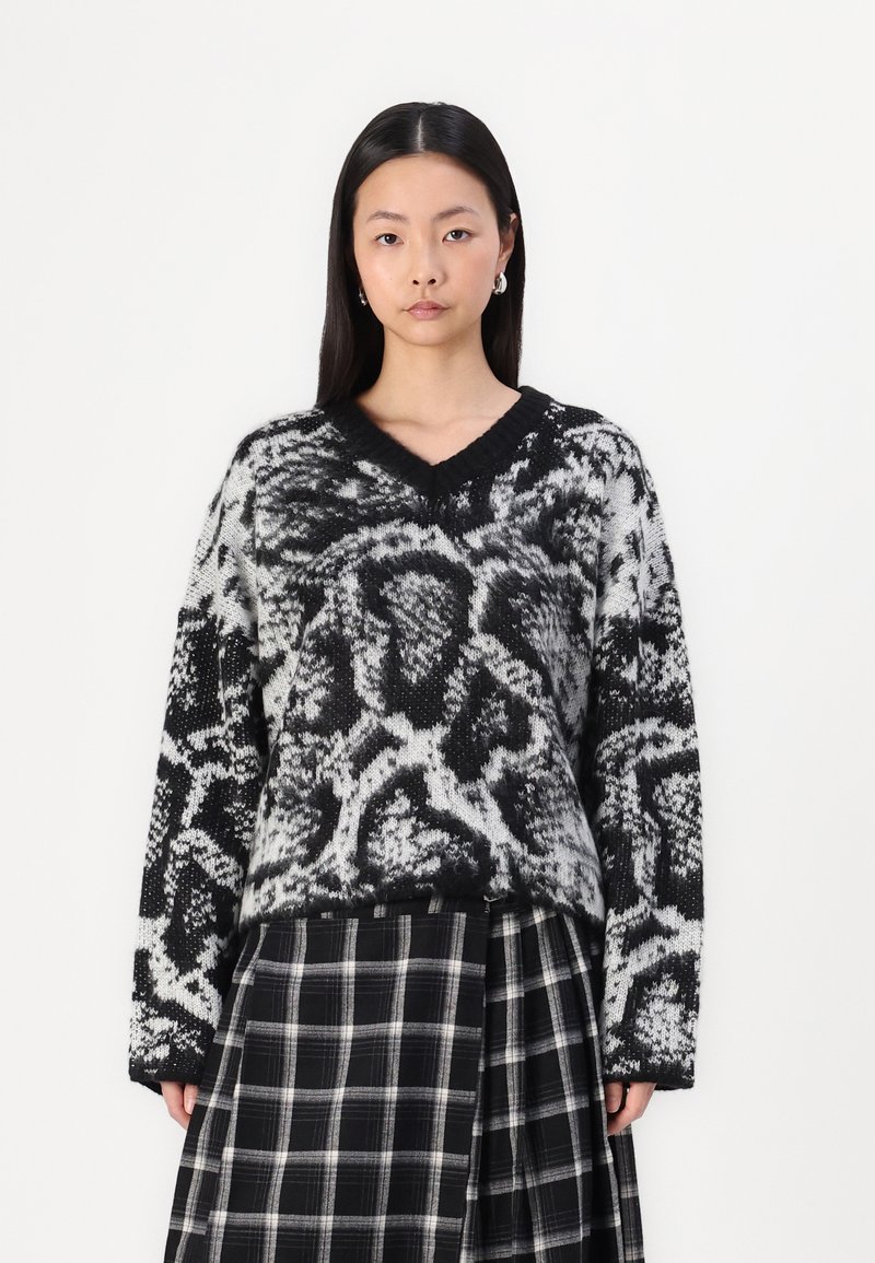 Maglione lavorato a maglia in bianco e nero con motivo, scollo a V, spalle scese e maniche lunghe, abbinato a una gonna a quadri in bianco e nero.