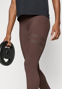 Καφέ κολάν με ομαλή υφή και διακριτικές ρίγες από δίχτυ στο επάνω μέρος του μηρού. Ένα άτομο κρατάει μια μαύρη πλάκα βάρους.