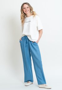 Bílá oversized tričko s textem "COOL MAMA", spárované s volnými světlomodrými kalhotami se stahovacími šňůrkami a tkanými bílými slip-on botami.