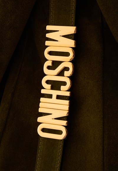 Μεταλλικά γράμματα σε χρυσό τόνο που σχηματίζουν τη λέξη «MOSCHINO», τοποθετημένα κάθετα σε φόντο από σκουροπράσινο σουέντ ύφασμα.