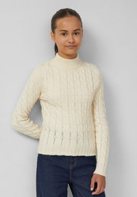 Strickpullover in Creme mit hohem Kragen, ausgestattet mit strukturierten Zopfmuster, geripptem Saum und langen Ärmeln. Kombiniert mit dunkelblauen Jeans.
