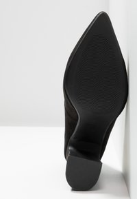 Chaussure à talon haut en daim noir avec bout pointu et semelle en caoutchouc noir texturée, présentant des rainures subtiles pour l'adhérence.