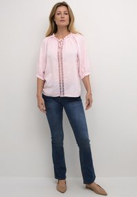 Helles rosa Leinenbluse mit gepufften Ärmeln und gerafftem Ausschnitt. Verfügt über Spitzenverzierungen an der Vorderseite. Kombiniert mit dunkelblauen Jeans.