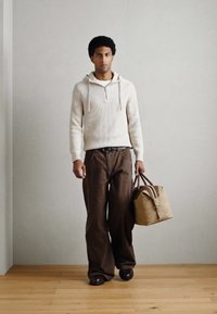 Camisola com capuz em malha de cor clara com fecho frontal, combinada com calças largas castanho-escuras. Acessório em pele castanha clara com duas alças.