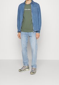 Kurtka dżinsowa, oliwkowa koszulka z logo "Calvin Klein", jasnoniebieskie dżinsy oraz wielokolorowe buty sportowe. Casualowy strój, mieszane materiały.