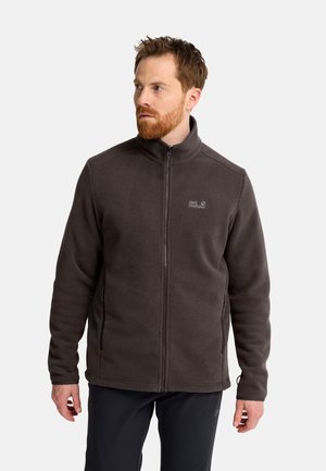 Mann mit roten Haaren und Bart, der eine dunkelbraune Fleecepullover mit Reißverschluss trägt, steht und schaut nach links auf einfarbigem Hintergrund.