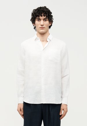 Jeune homme aux cheveux bouclés portant une chemise blanche à manches longues boutonnée et un pantalon foncé, debout devant un fond uni.