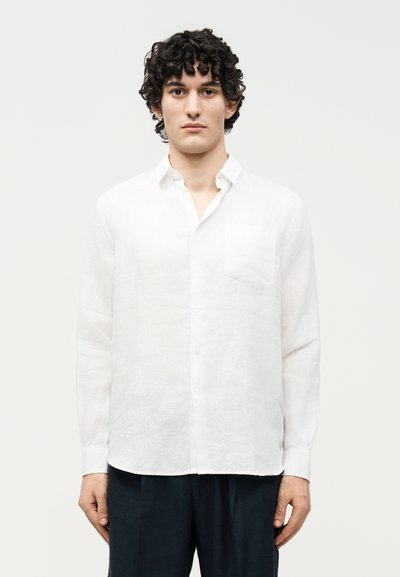 Jeune homme aux cheveux bouclés portant une chemise blanche à manches longues boutonnée et un pantalon foncé, debout devant un fond uni.